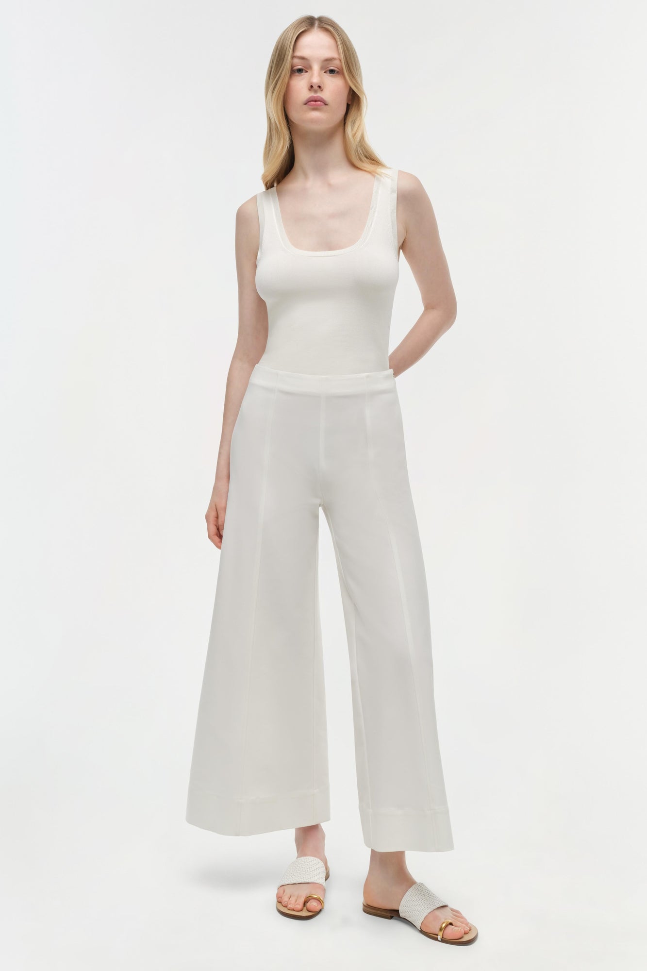 Woman wearing off-white sleeveless tank top and cropped wide-leg cotton pants in cream, paired with white slides.