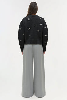 Black knit crewneck sweater with scattered silver embellishments worn with gray wide-leg pants, shown from back view.