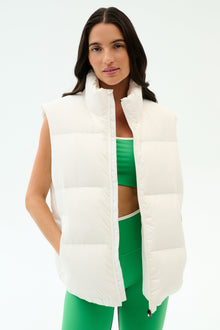 A woman models the SPLITS59 Summit Nylon Puffer Vest in white over a green sports bra and matching leggings, standing against a plain white backgroundâ€”ideal for lightweight insulation during cool weather workouts.