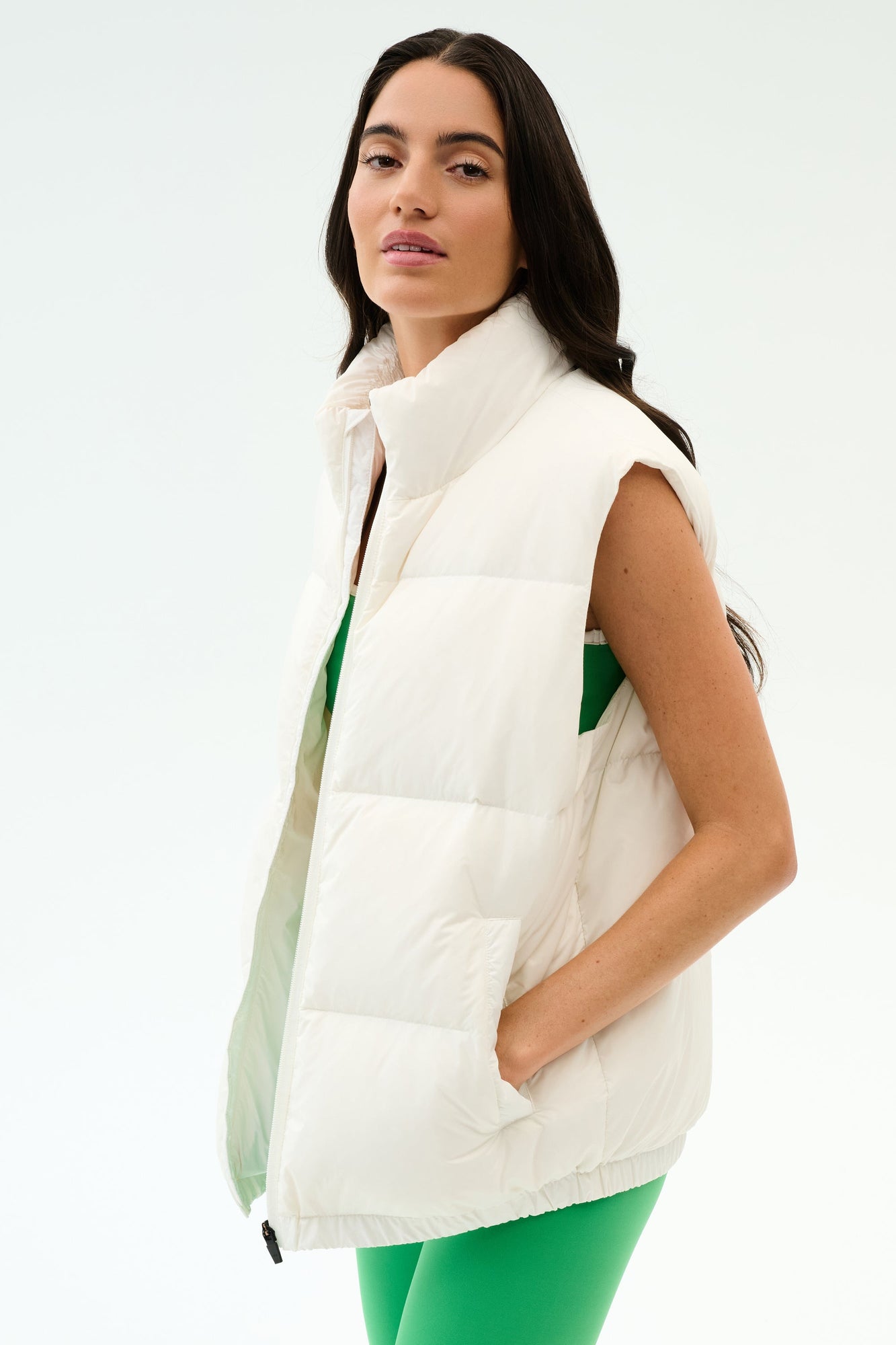 A woman sports the SPLITS59 Summit Nylon Puffer Vest in white over a green outfit, standing with one hand in her pocket against a plain backdropâ€”perfect lightweight insulation for cool weather workouts.