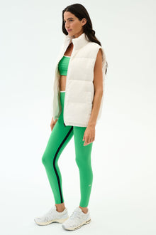A woman stands in profile against a plain background wearing the SPLITS59 Summit Nylon Puffer Vest in white, paired with a green sports bra, green leggings with black stripes, and white athletic shoesâ€”ideal for cool weather workouts.