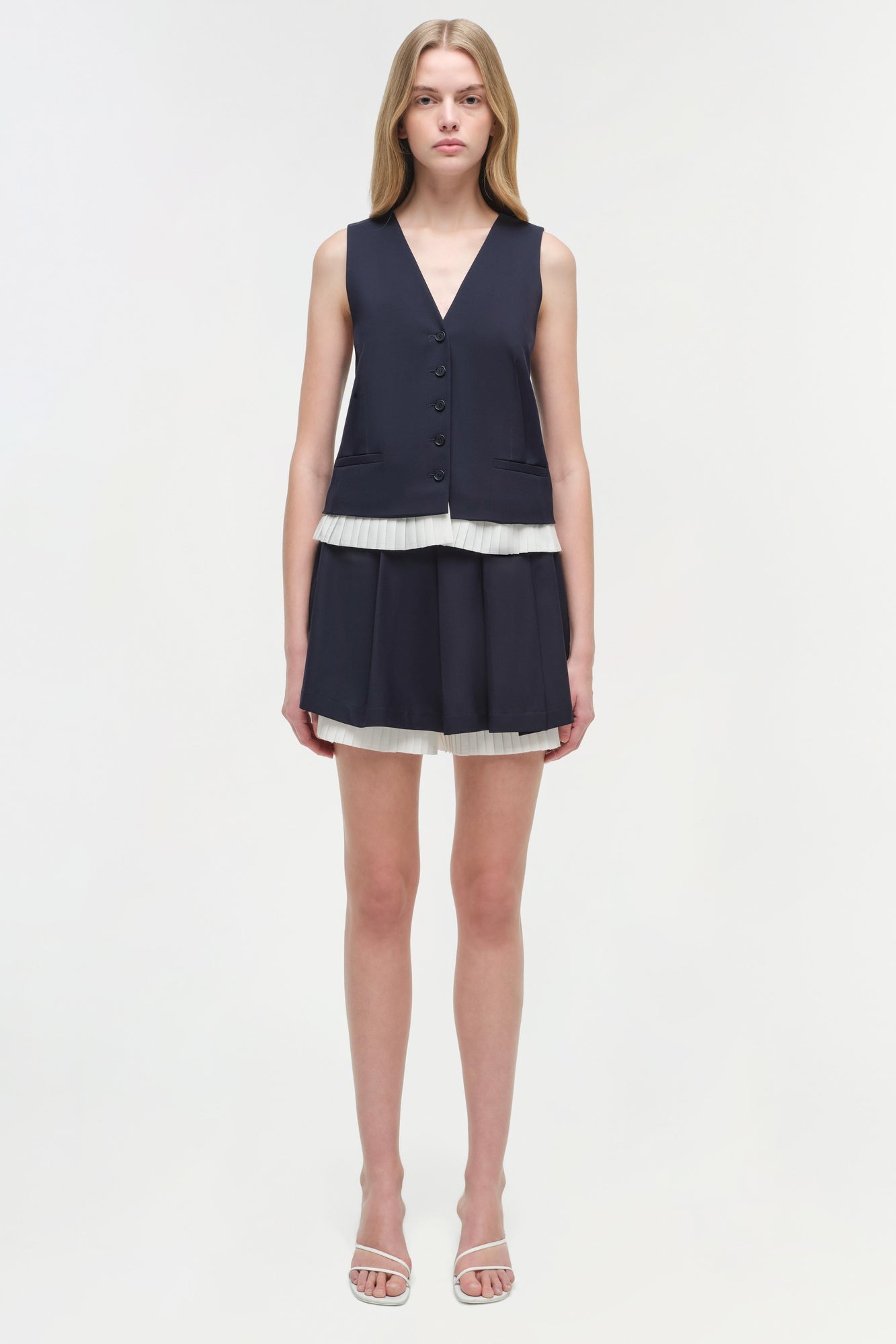 Woman wearing navy sleeveless button-front vest with pleated skort featuring white trim and matching white trim at waist.
