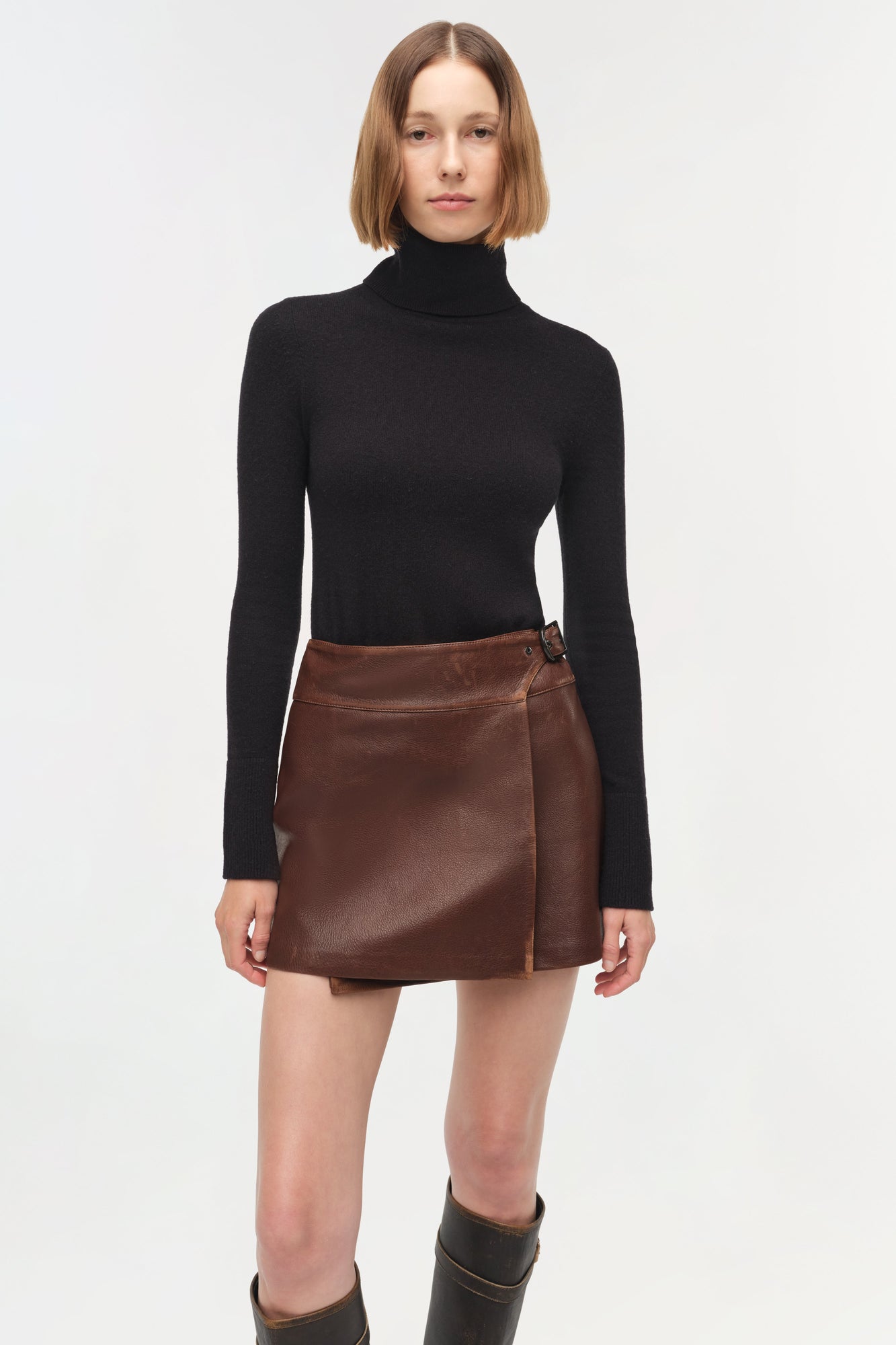 Brown leather mini skirt with wrap-front closure and side button detail, worn with black turtleneck.