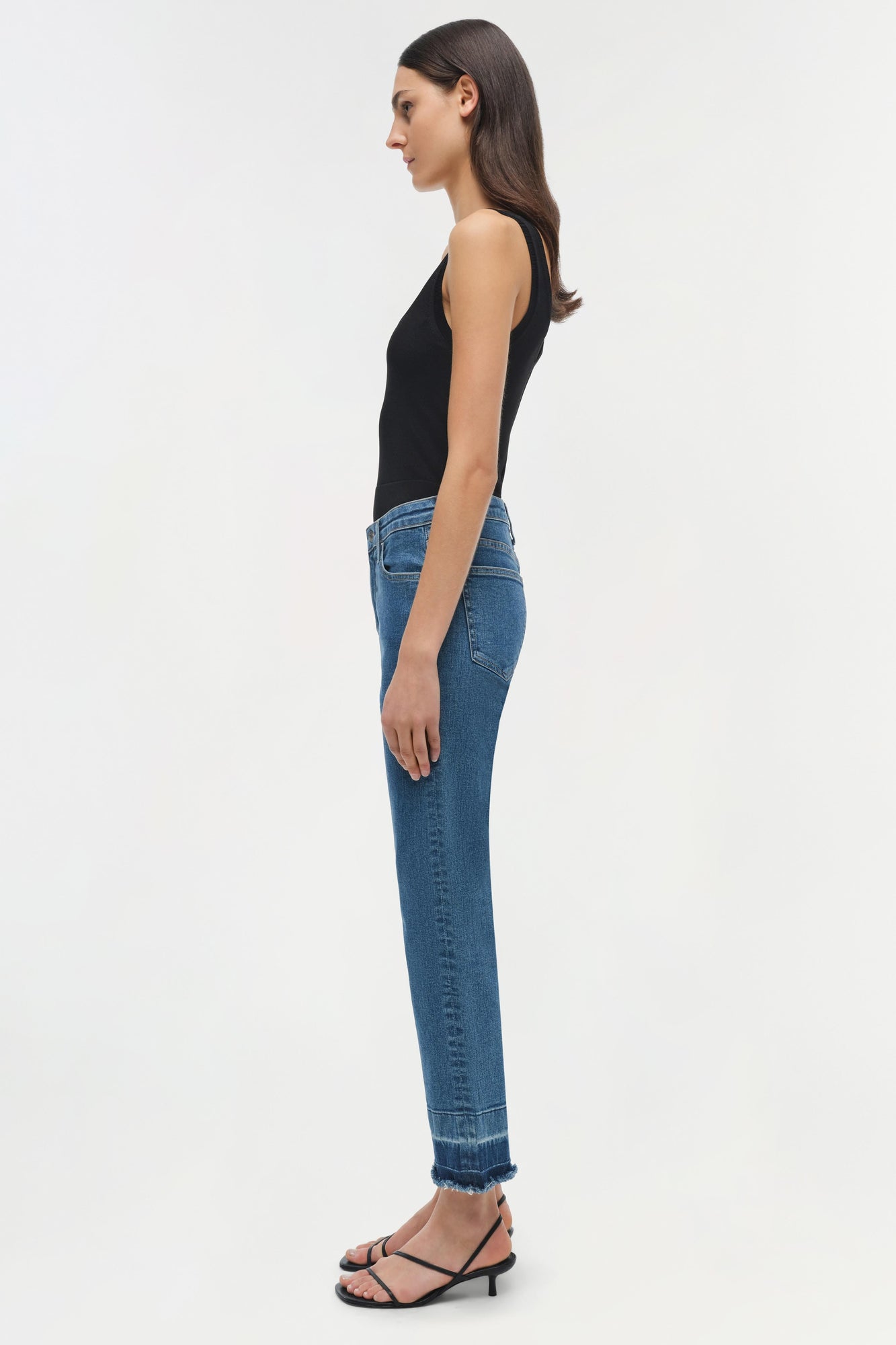 Woman wearing black sleeveless tank top and blue straight-leg jeans with frayed ankle hem, shown in side profile.