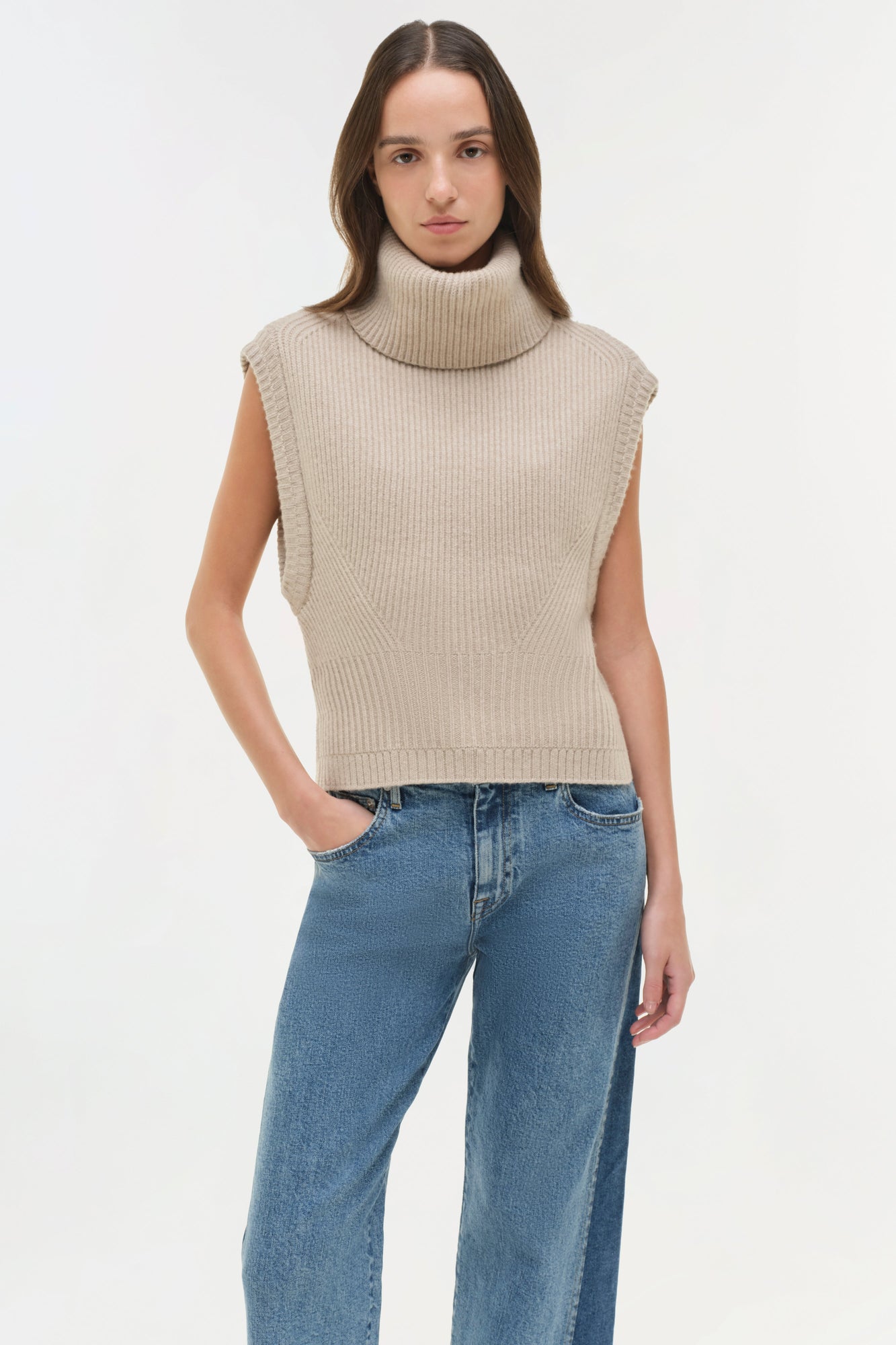 Woman wearing beige sleeveless turtleneck sweater and blue straight-leg jeans.