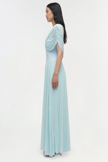 Light blue jersey maxi dress with draped cape overlay and pleated skirt, shown on model in profile view.