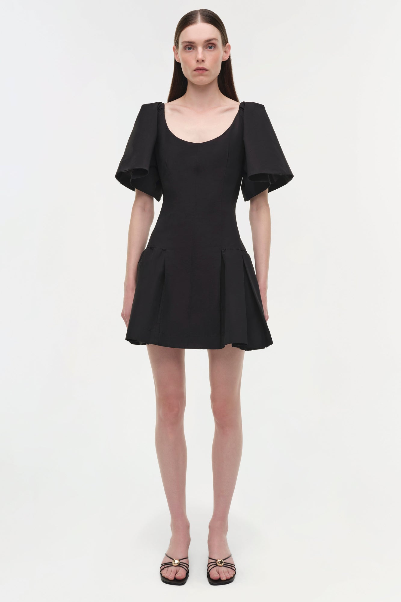 Black mini dress with puffed short sleeves, scoop neckline, and pleated skirt, worn with black strappy sandals.