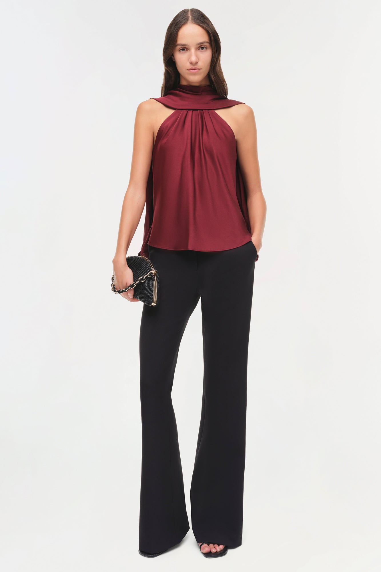 Woman wearing burgundy sleeveless halter top with draped cape detail and black flared pants.