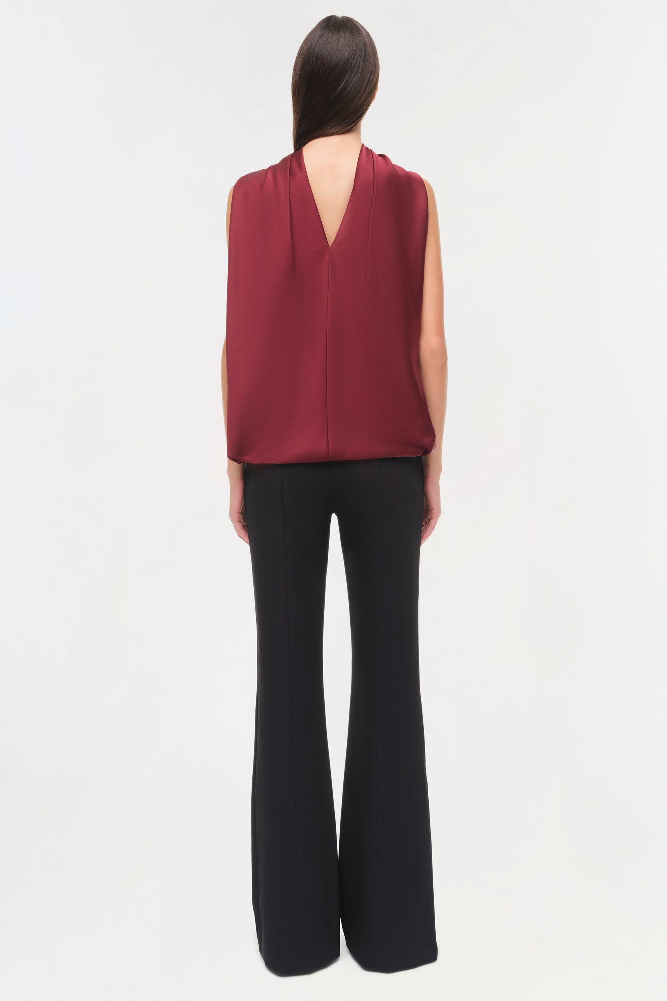 Woman wearing burgundy sleeveless satin cape top with draped back over black flared pants, shown from behind.