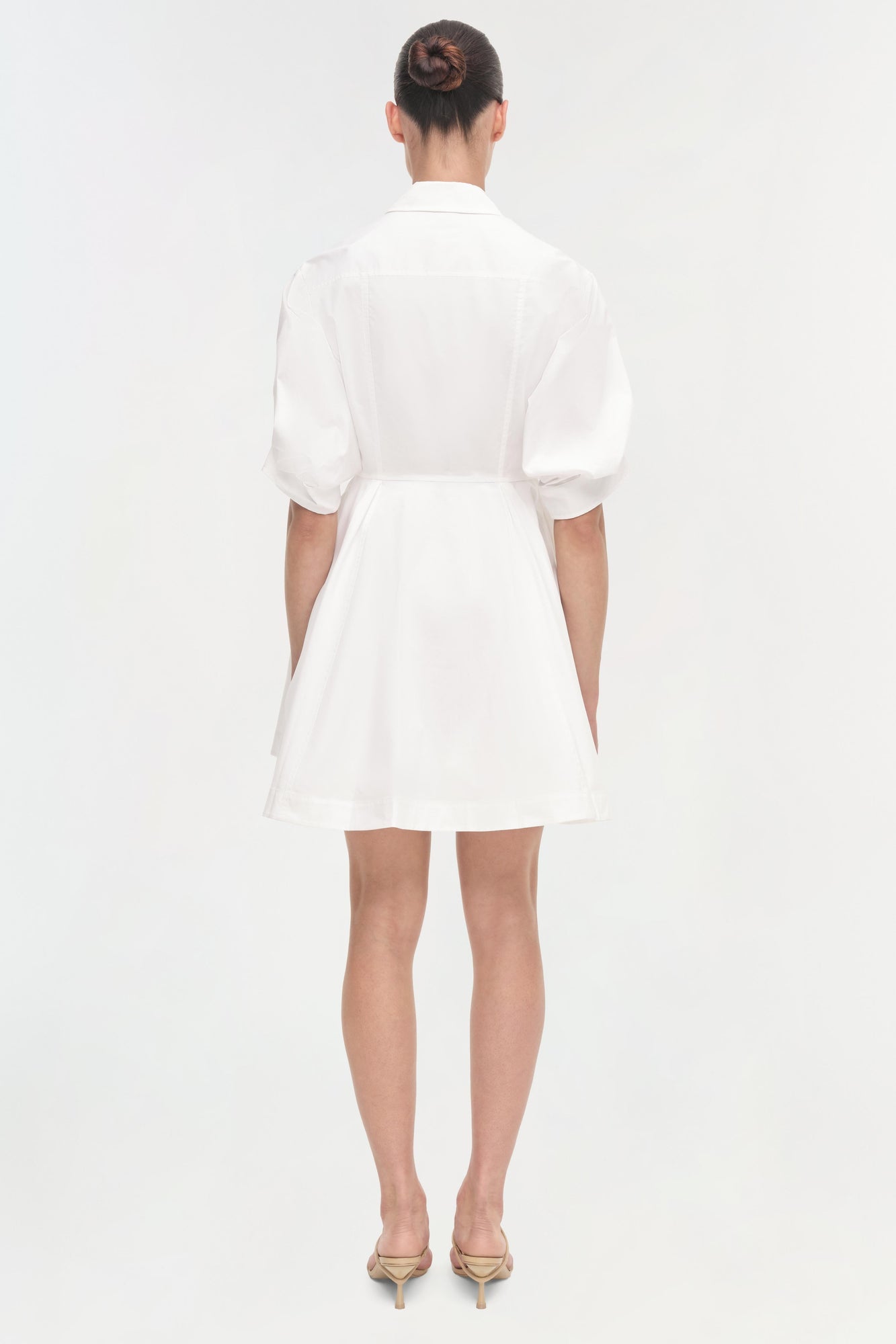 Woman wearing white collared dress with puffed sleeves and belted waist, shown from back view, with nude heels.