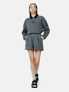 A female model wearing a black oversized French terry crew sweatshirt with a relaxed drop shoulder silhouette, paired with shorts and black sandals.
