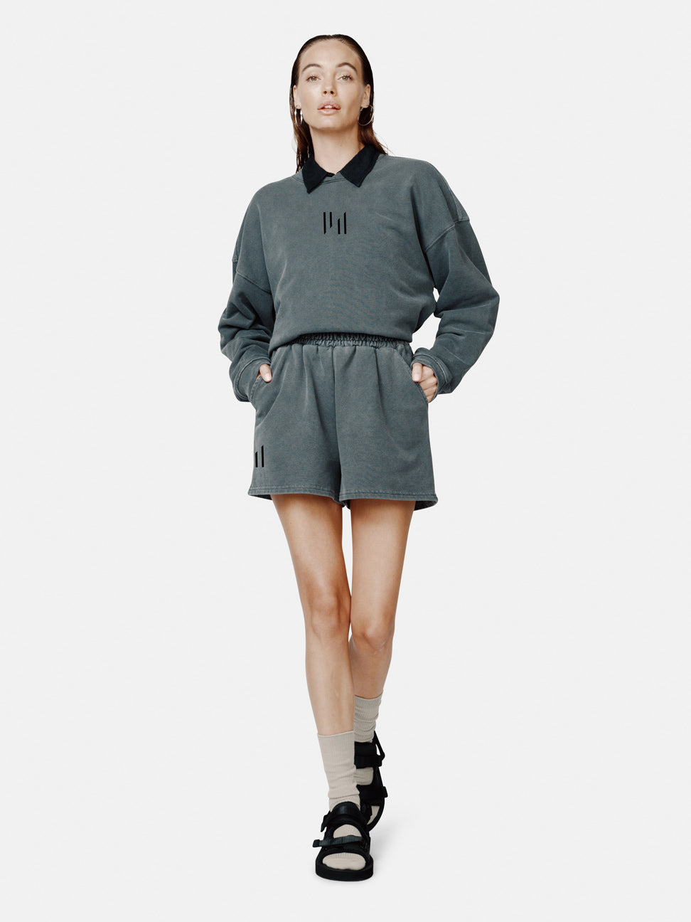 A female model wearing a black oversized French terry crew sweatshirt with a relaxed drop shoulder silhouette, paired with shorts and black sandals.