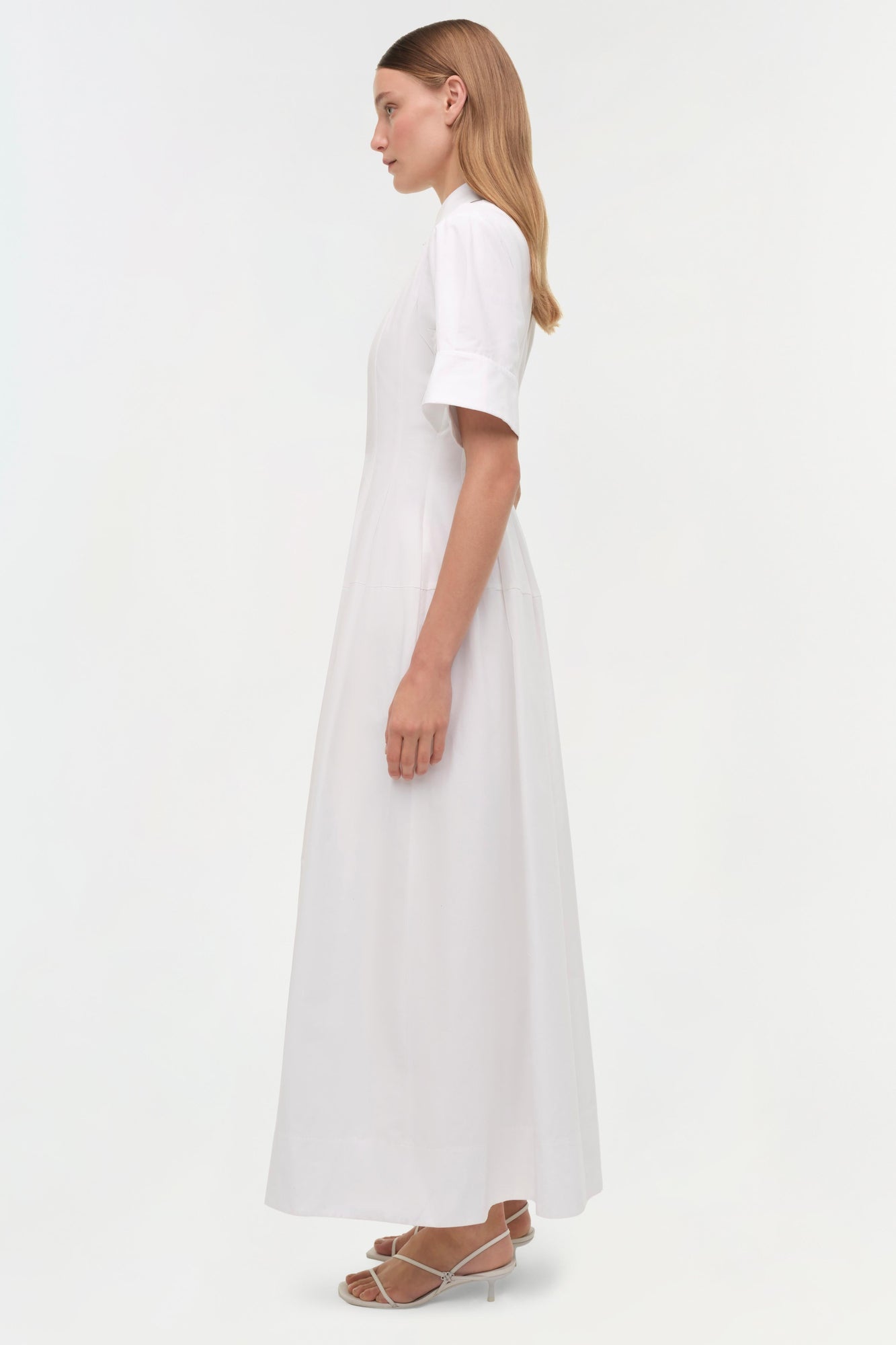 Woman wearing a white cotton shirt dress with short sleeves and an A-line skirt, shown in profile view.