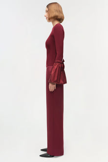 Woman wearing burgundy knit top with taffeta ruffle detail at waist, shown in profile view.