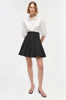 Woman wearing white collared button-up shirt with three-quarter sleeves and black pleated mini skirt with side pockets.