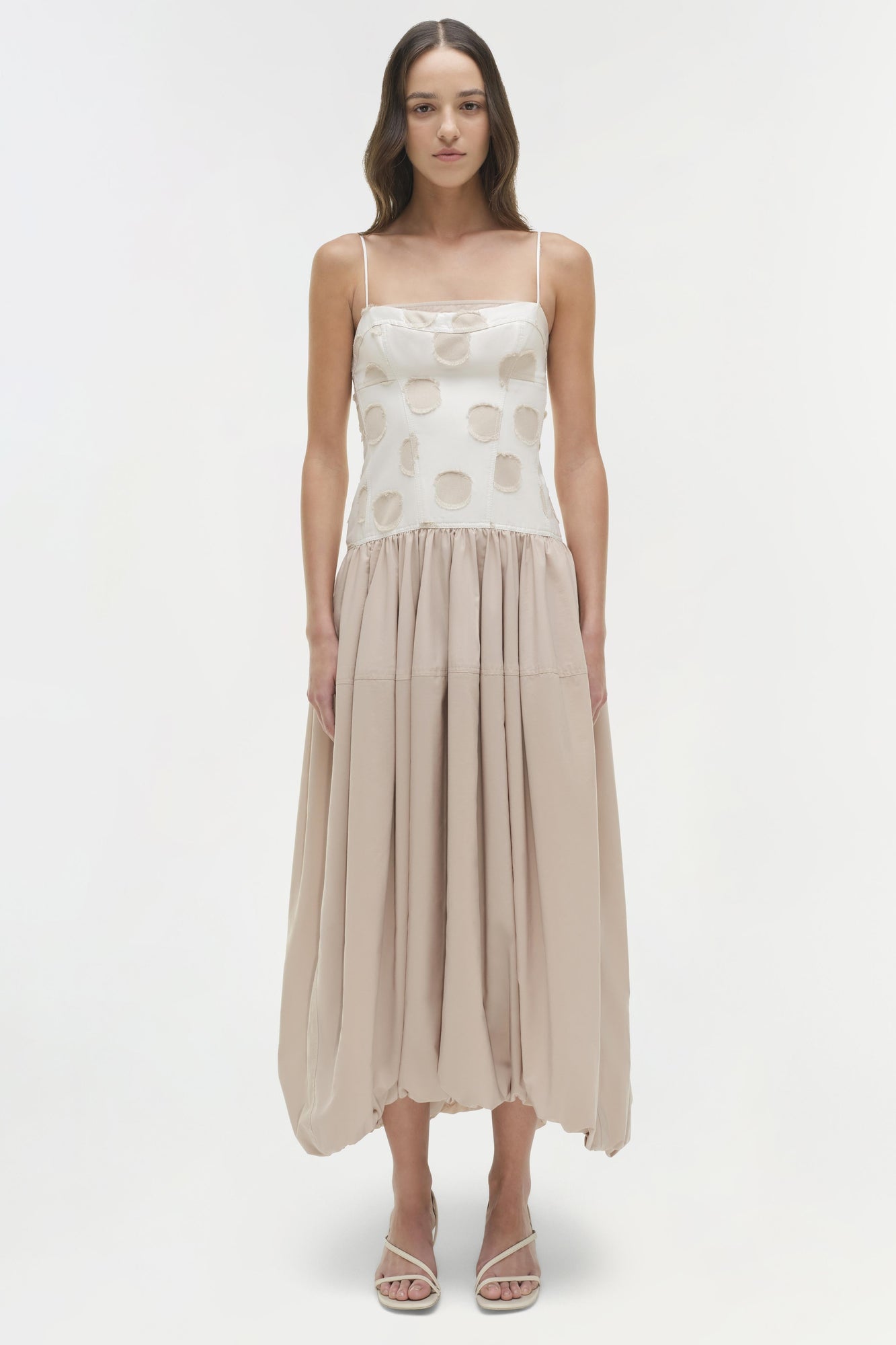 Woman wearing a cream and beige midi dress with geometric circle print top and pleated skirt, paired with white sandals.