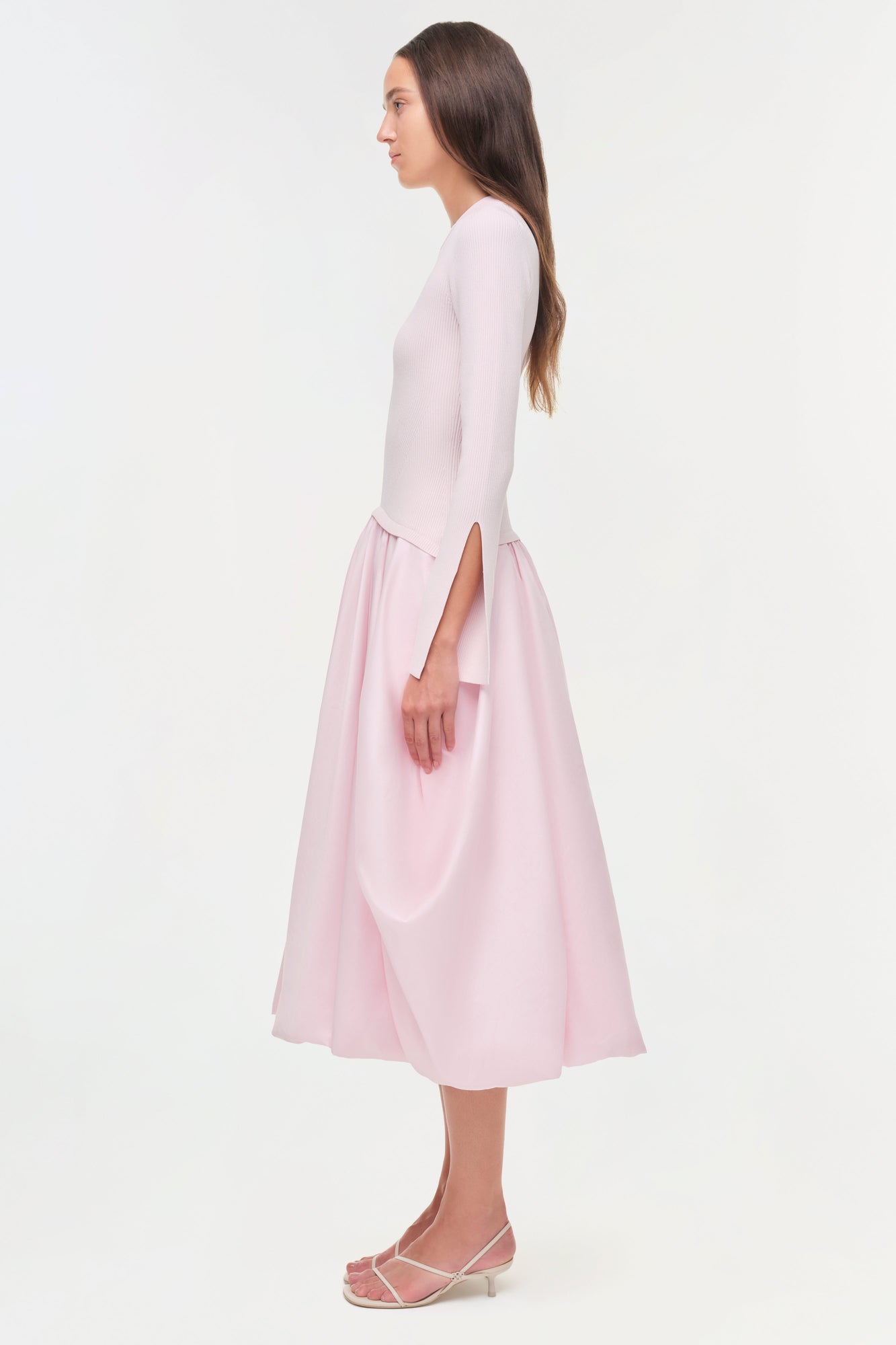 Woman wearing a light pink midi dress with a fitted ribbed knit top and flowing taffeta skirt, shown in profile view.