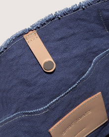 Close-up of a blue textile bag by graf lantz, made from durable cotton canvas with frayed edges. Inside, the Hana Canvas Tote features a tan leather strap with black snap button and textured tan patch with visible stitches. The fabric looks soft and sturdy.