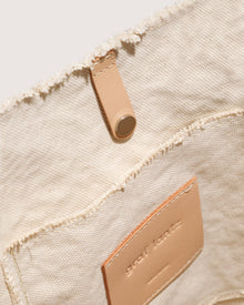 Close-up interior of a graf lantz beige fabric bag with a small leather logo patch and leather tab featuring a metal snap button. The textured cotton canvas, with frayed edges, embodies the signature craftsmanship of the Hana Canvas Tote.