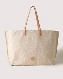 The graf lantz tote bag is a minimalist, large beige canvas with tan vegetable-tanned leather straps and a small square leather patch on the front, set against a simple light background.