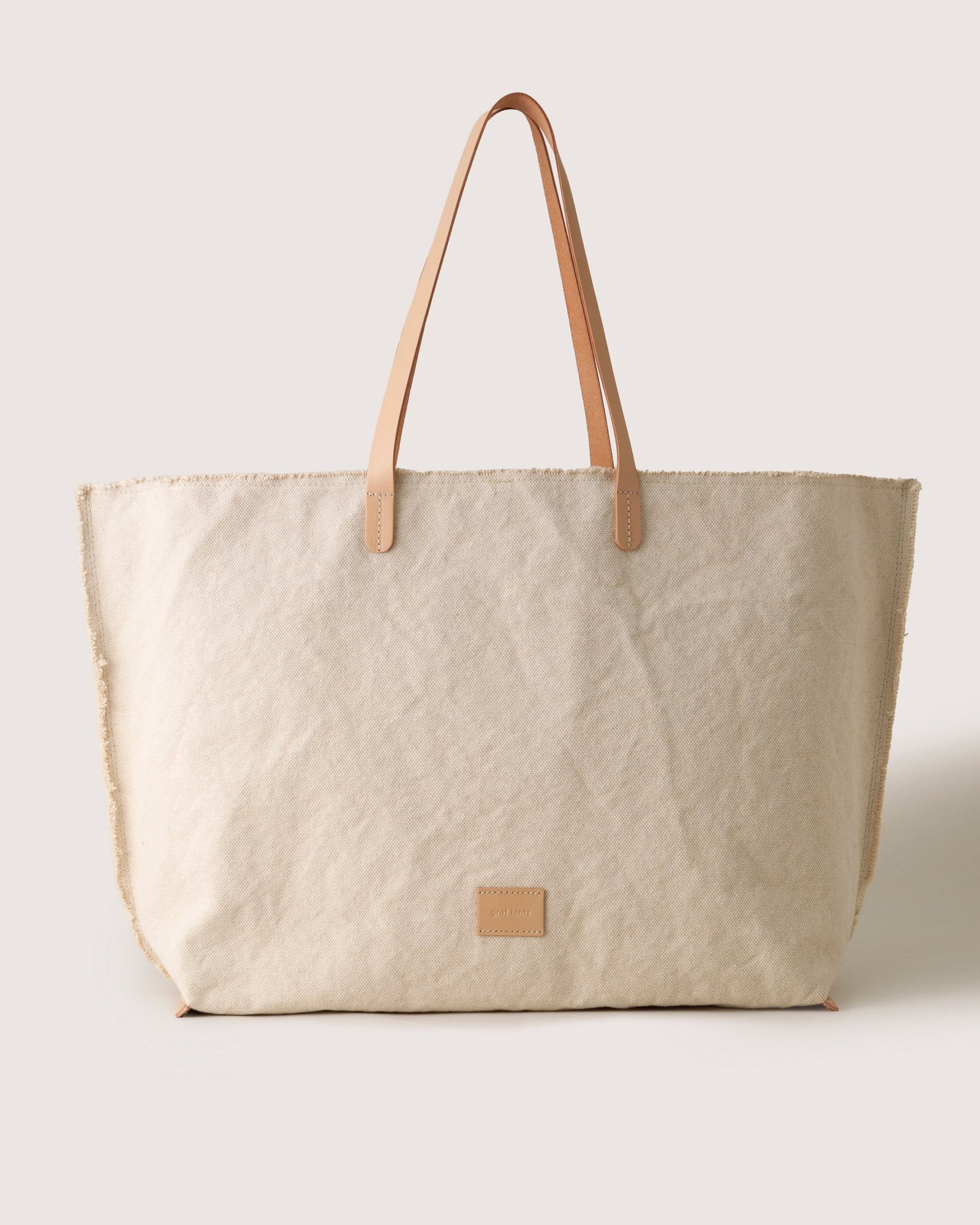 The graf lantz tote bag is a minimalist, large beige canvas with tan vegetable-tanned leather straps and a small square leather patch on the front, set against a simple light background.