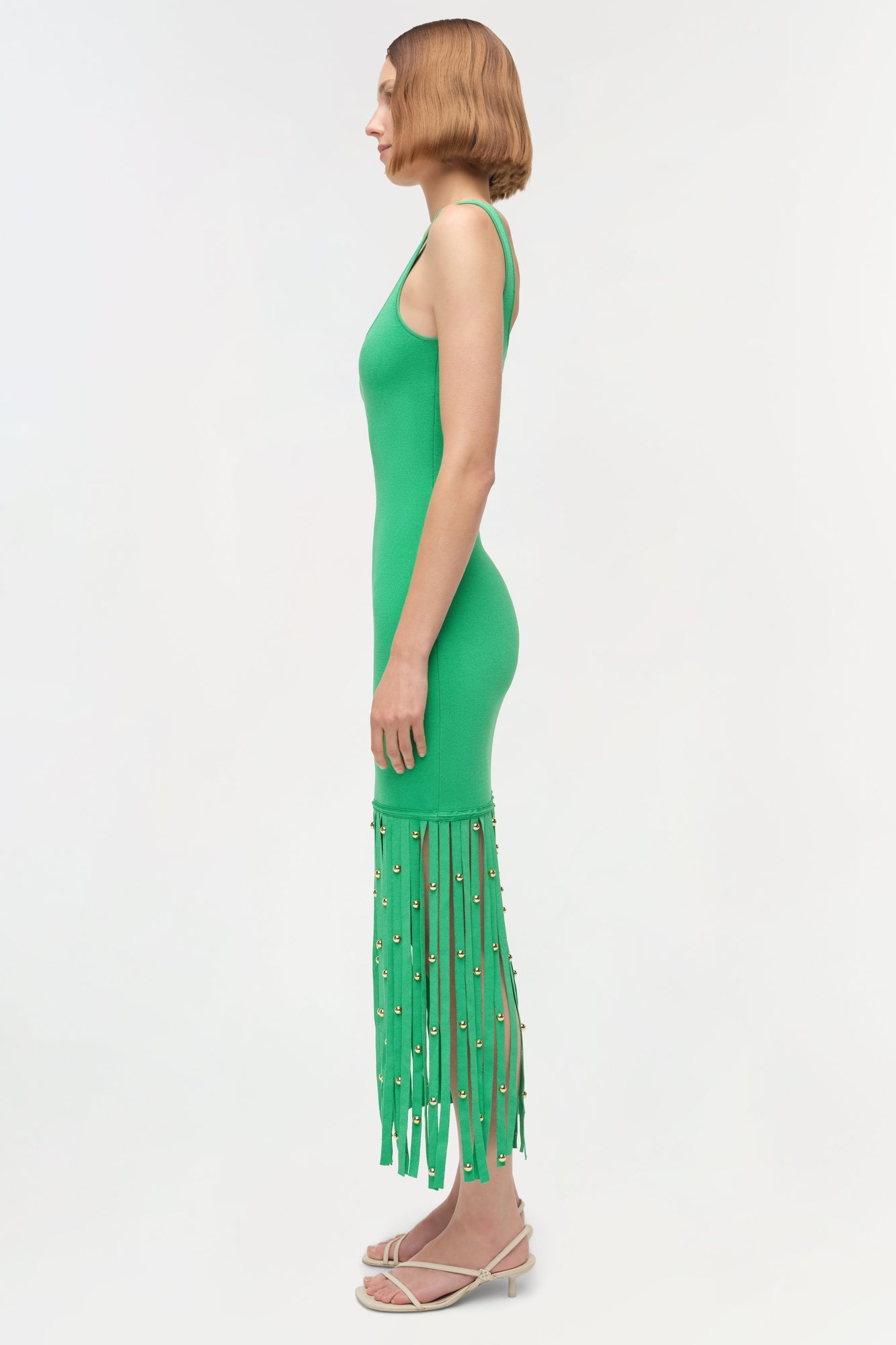 Woman wearing sleeveless green knit midi dress with beaded fringe hem detail, shown in profile view.