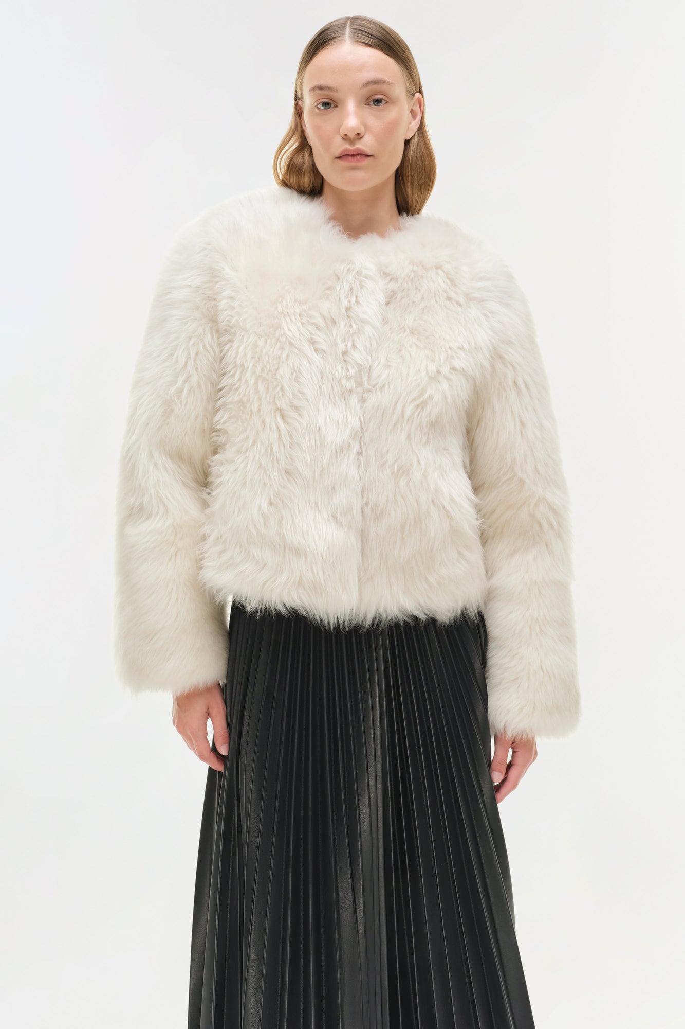 Cream shearling jacket with fluffy texture and cropped length, worn with a black pleated skirt.