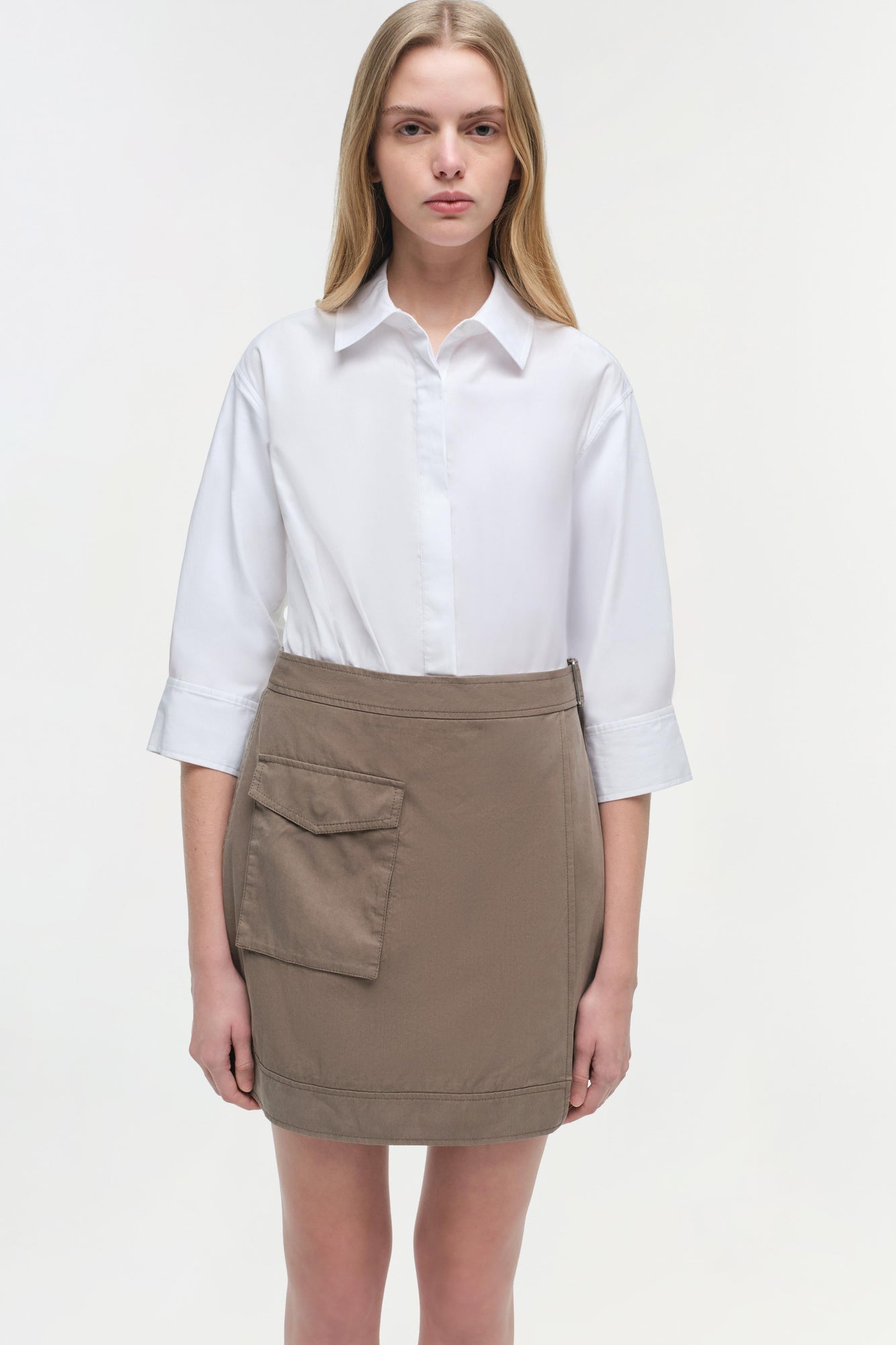 Woman wearing white button-up shirt with three-quarter sleeves and taupe mini skirt with cargo pocket detail.