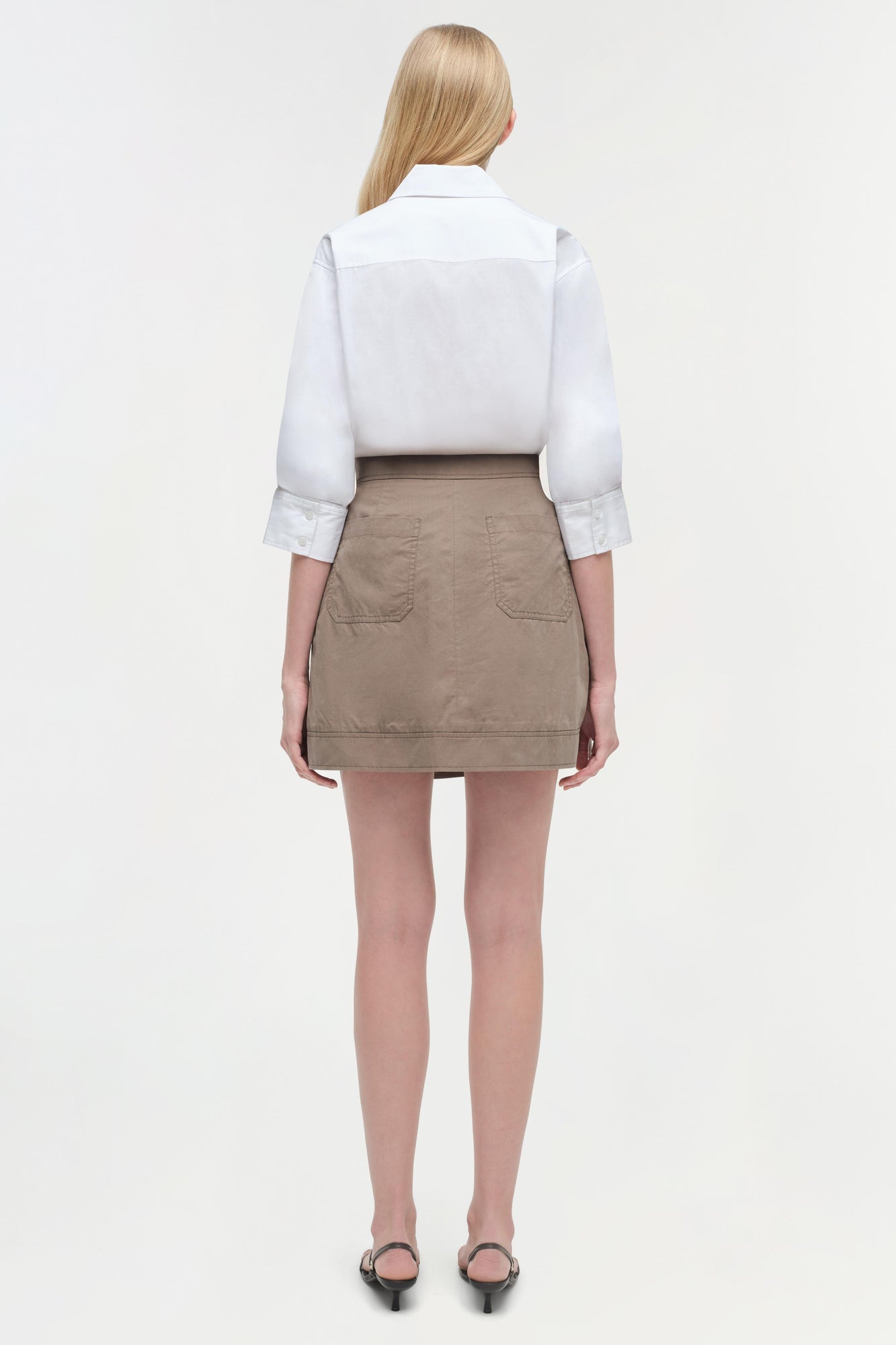 Model wearing white collared shirt with three-quarter sleeves over beige mini skirt with patch pockets, viewed from back.