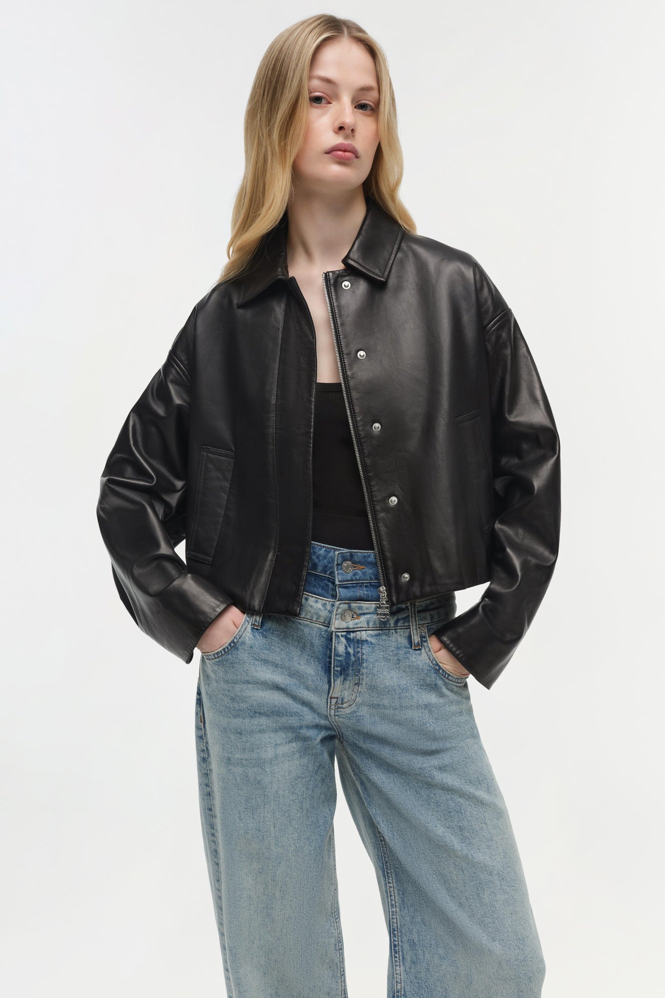 Black leather jacket with collar, front button closure, and chest pockets worn over black top and blue jeans.