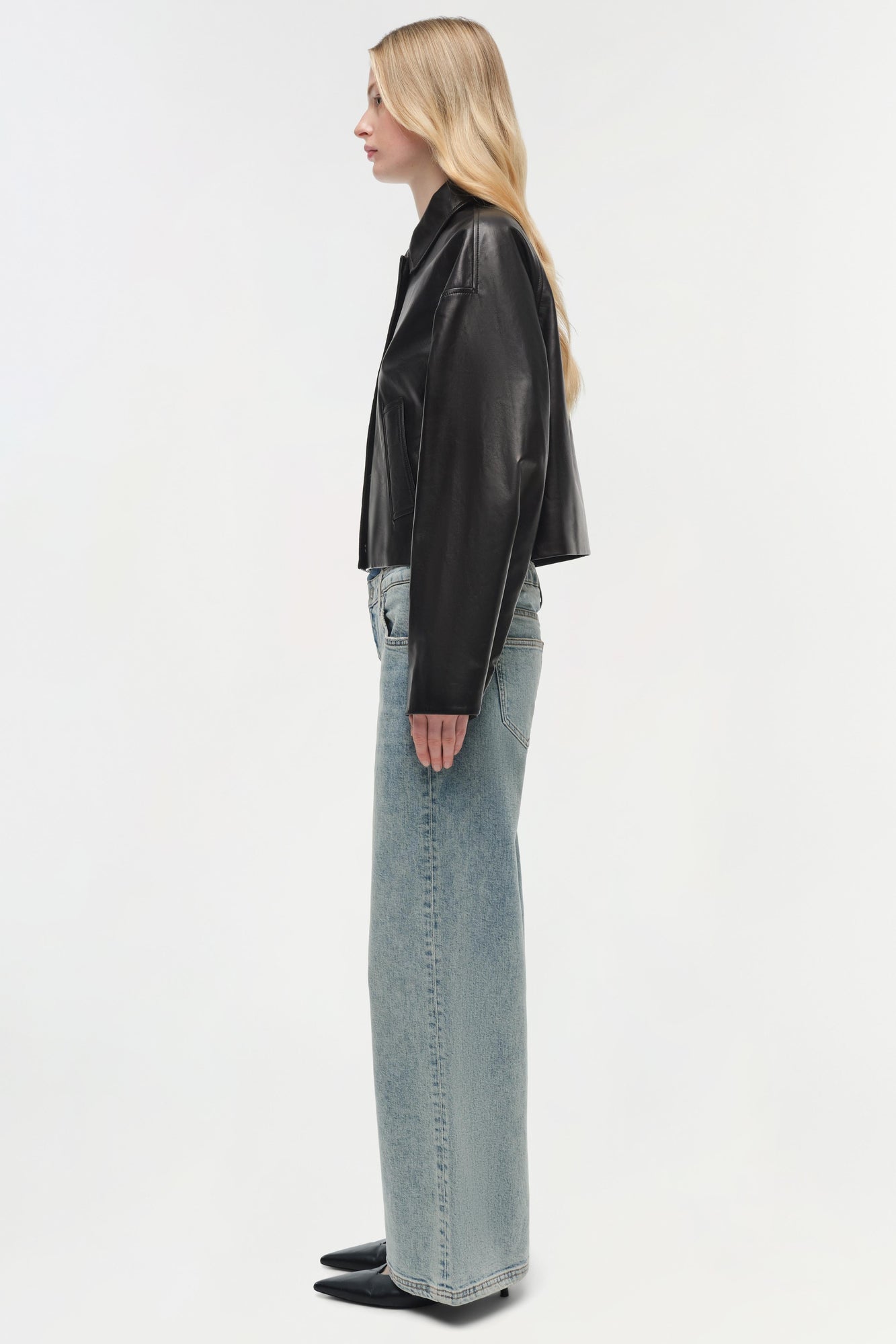 Woman wearing black leather jacket with long sleeves and light-wash denim jeans, shown from side profile.