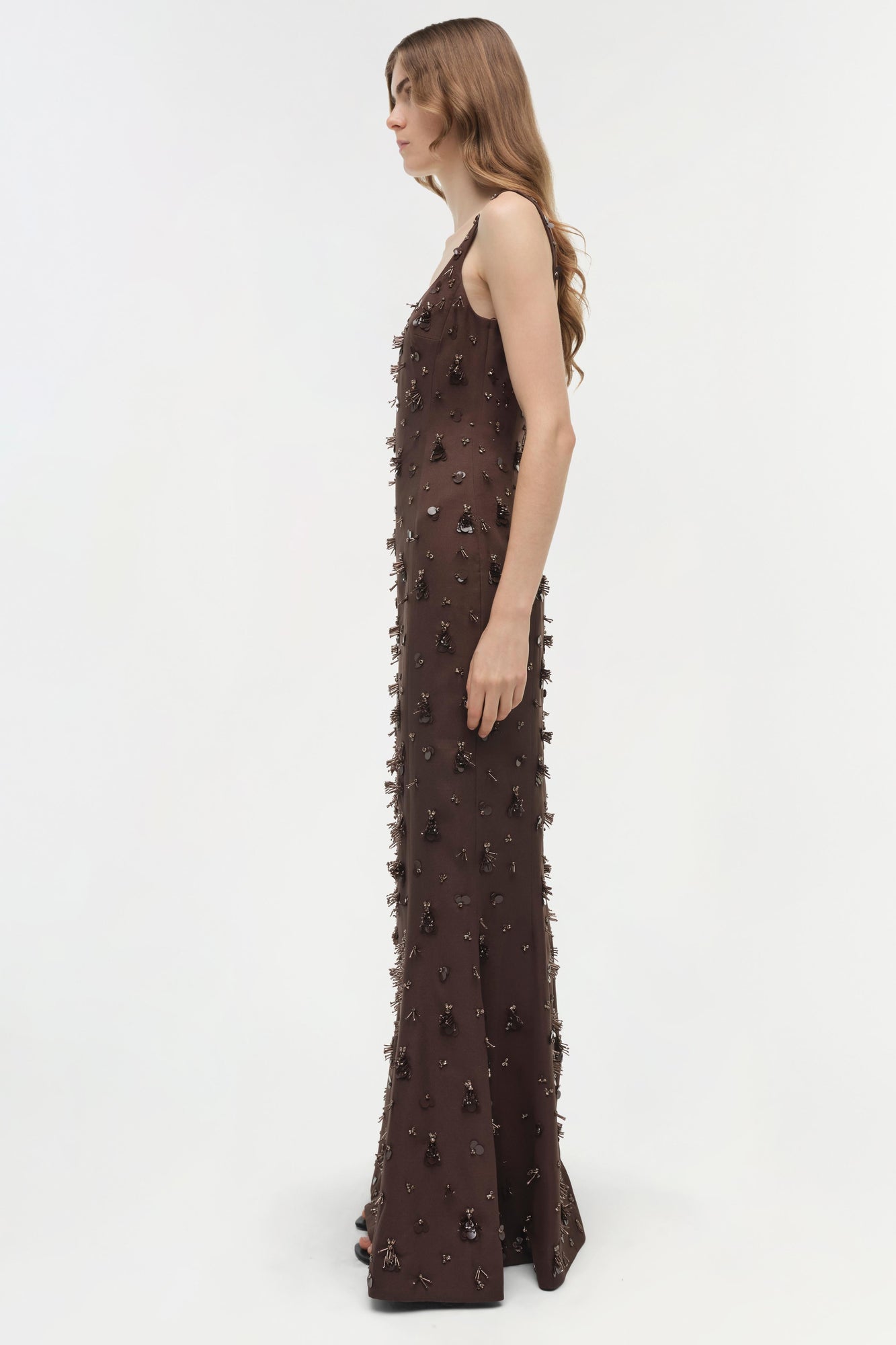 Brown sleeveless floor-length gown with beaded and fringe embellishments throughout, shown on model in profile.