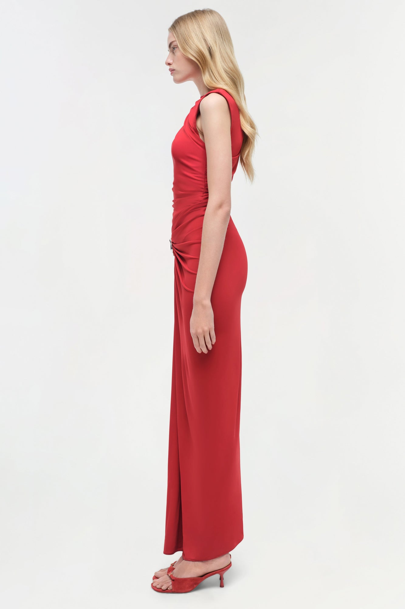Woman wearing a red sleeveless jersey gown with ruched side detailing and matching heeled sandals.