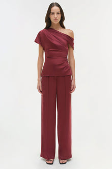 Burgundy one-shoulder draped satin top with short sleeve and ruched waist detail, worn with matching wide-leg pants.
