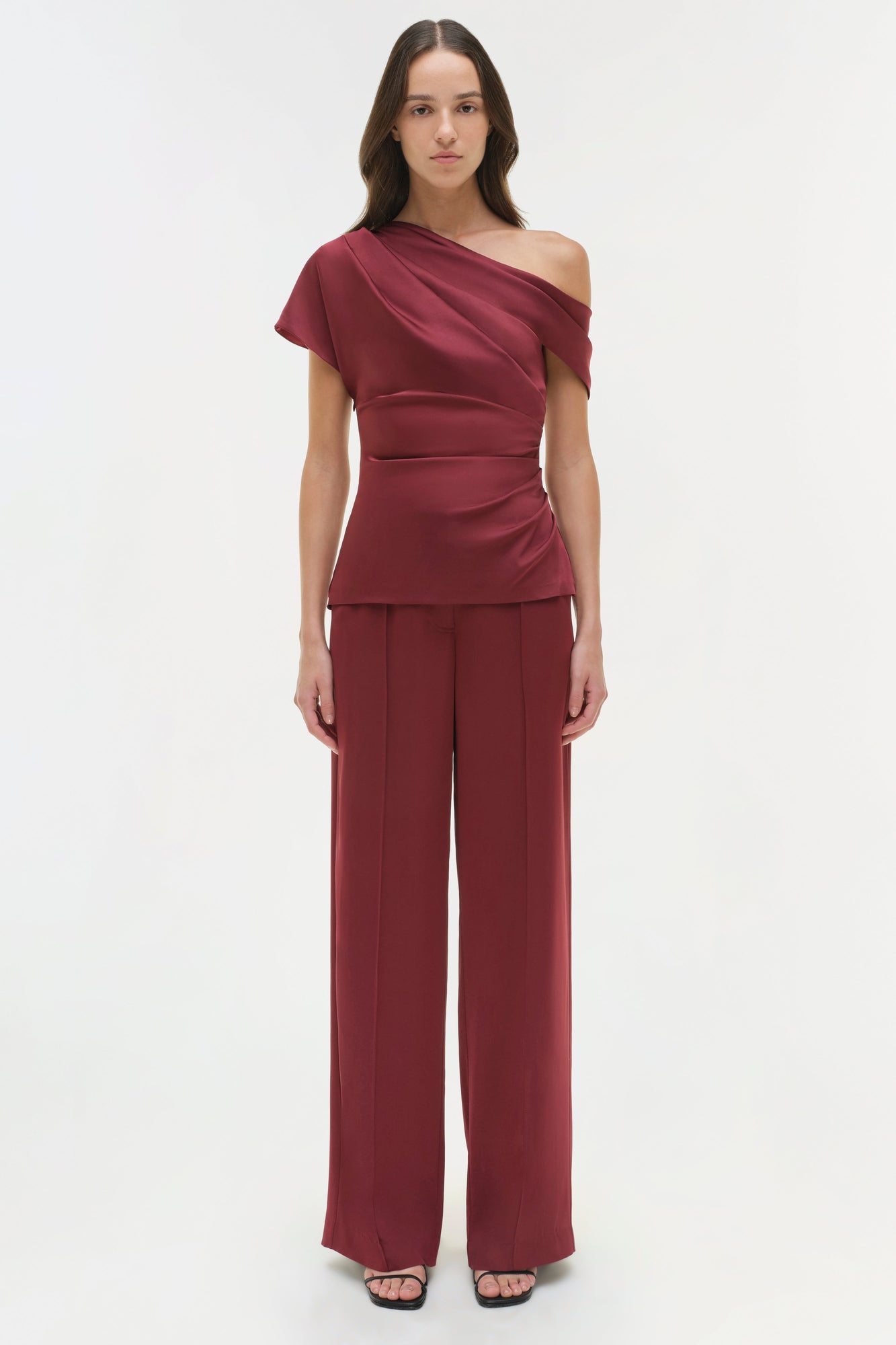 Burgundy one-shoulder draped satin top with short sleeve and ruched waist detail, worn with matching wide-leg pants.
