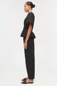 Woman wearing black pull-on pants with a peplum top, shown in profile view.