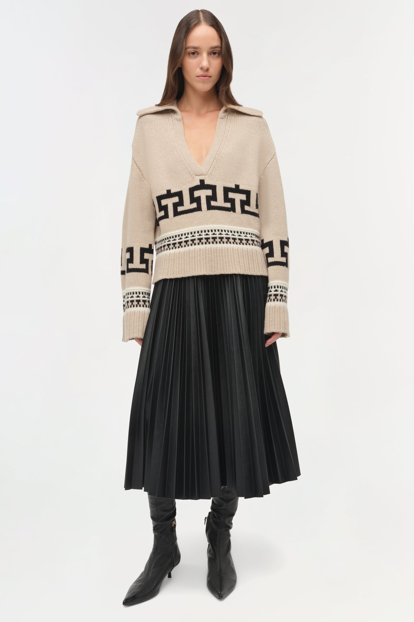 Beige V-neck sweater with black geometric and geometric patterns worn with black pleated skirt and boots.