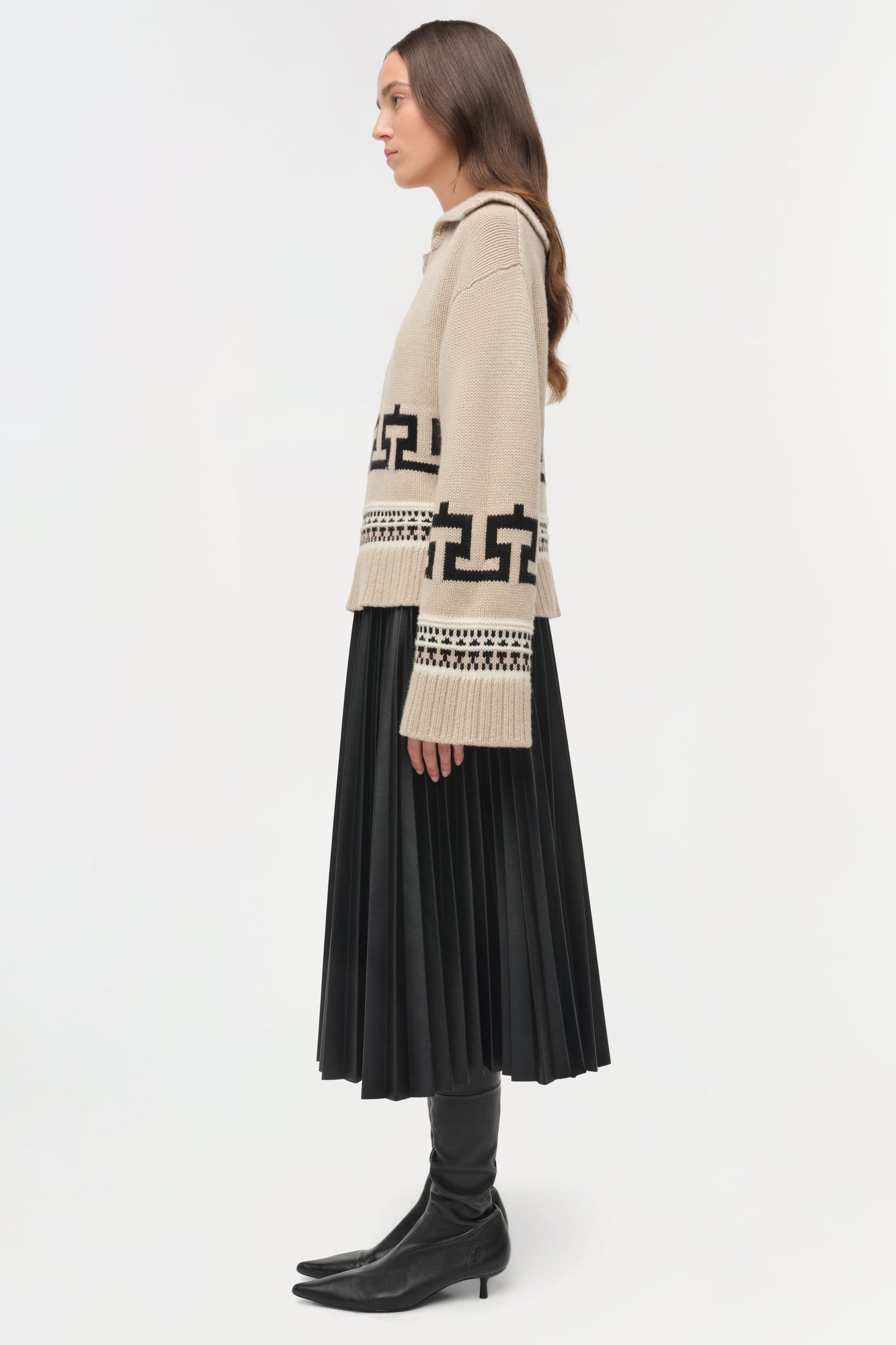 Cream V-neck sweater with black geometric patterns worn over black pleated skirt and boots.