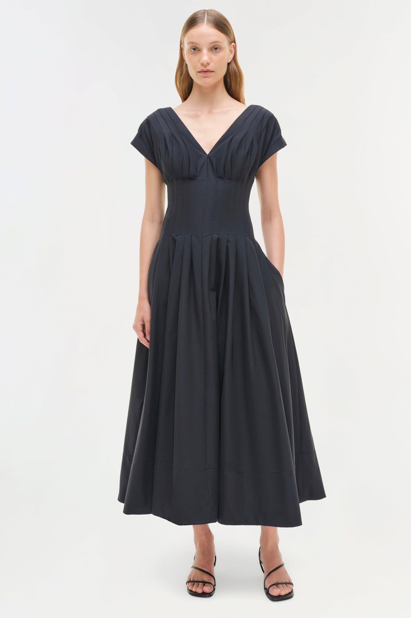 Black cotton midi dress with short sleeves, V-neckline, fitted waist, and full pleated skirt, modeled on woman.
