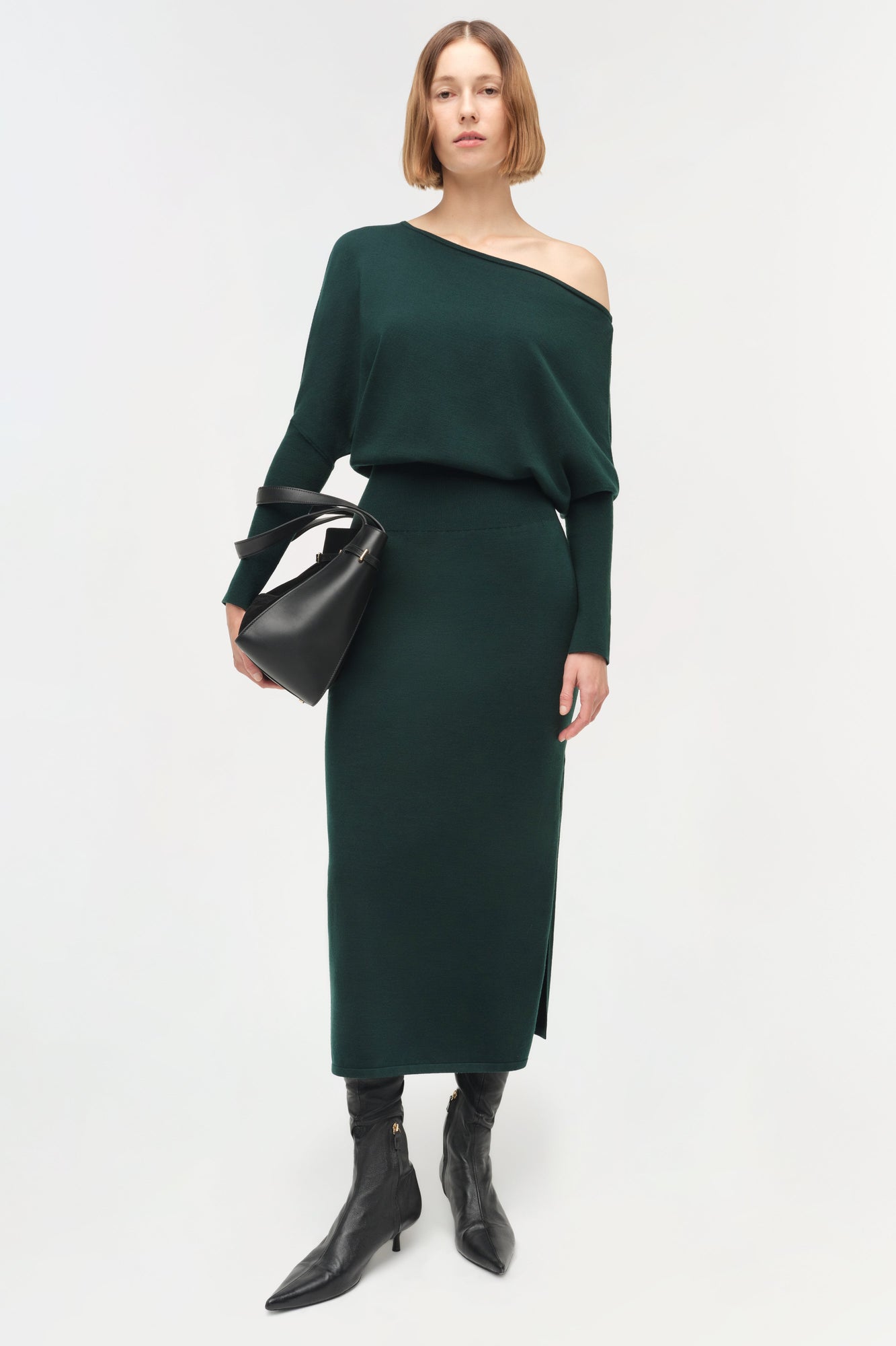 Woman wearing dark green off-shoulder midi dress with long sleeves and a fitted waist, paired with black boots and a black ba