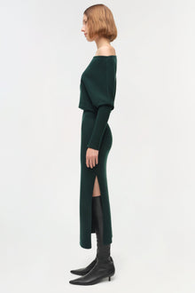 Woman wearing dark green off-shoulder midi dress with long sleeves and front slit, styled with black boots.