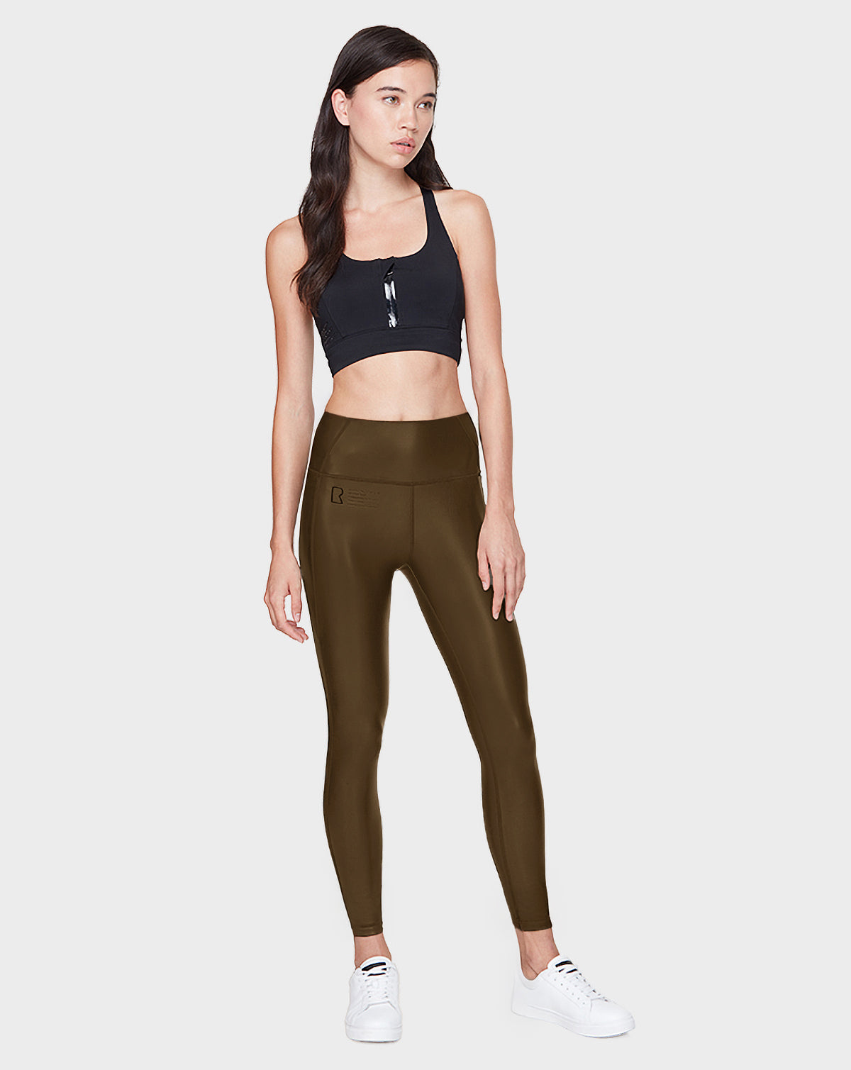 A woman poses against a light grey backdrop wearing the SHIRIN L - 8521034 Army high-waisted olive green leggings by Rudsak, paired with a black sports bra and white sneakers. She has long brown hair and a neutral expression.