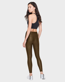 A woman poses confidently against a white background, wearing the Rudsak SHIRIN L - 8521034 Army vegan leather leggings with a black crop top and silver sneakers, glancing back over her shoulder.
