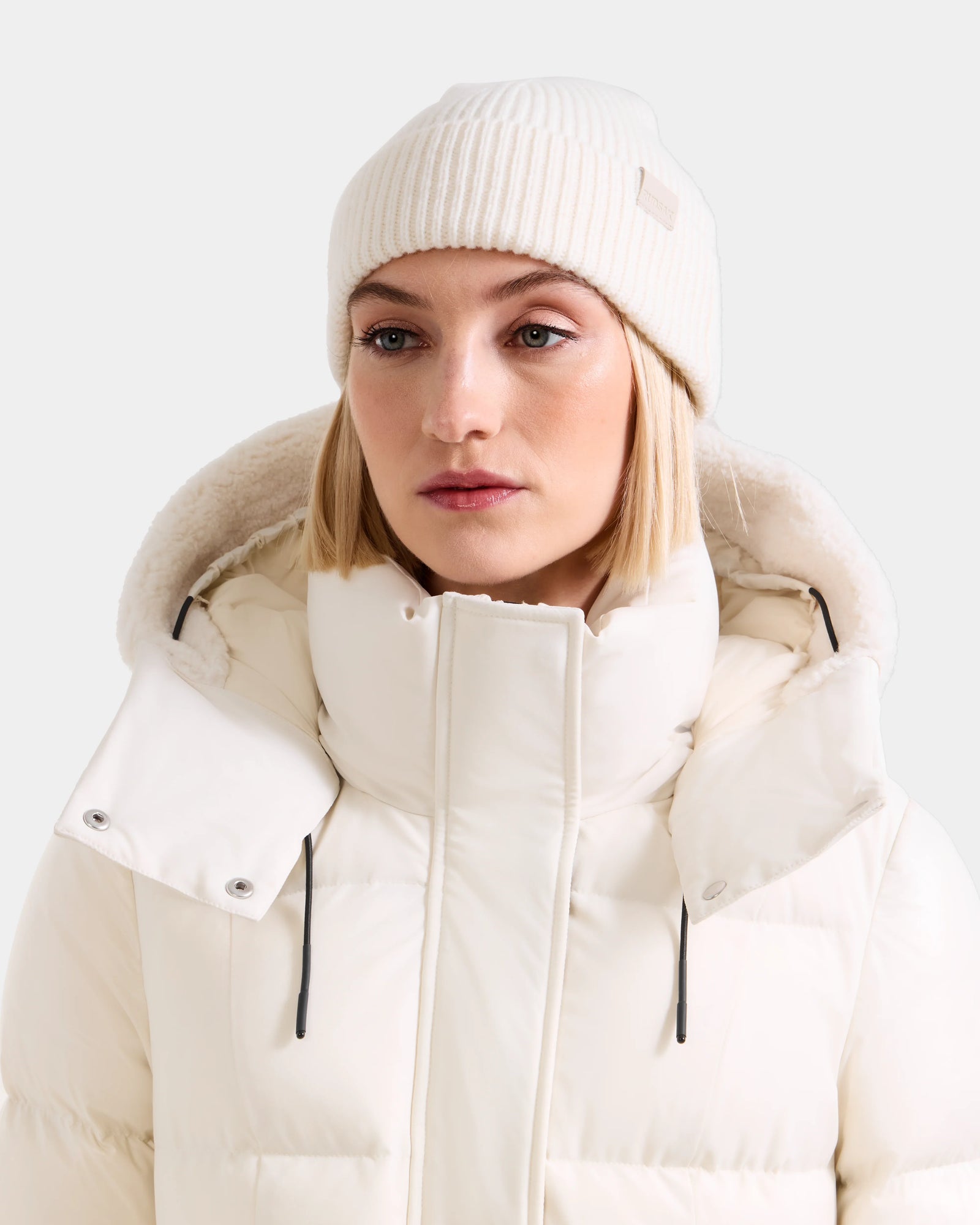 A woman wears the Rudsak ANDREY - U425507 Vanilla rib-knit hat and thick white puffer jacket with high collar and hood, looking slightly to the side against a plain light background.