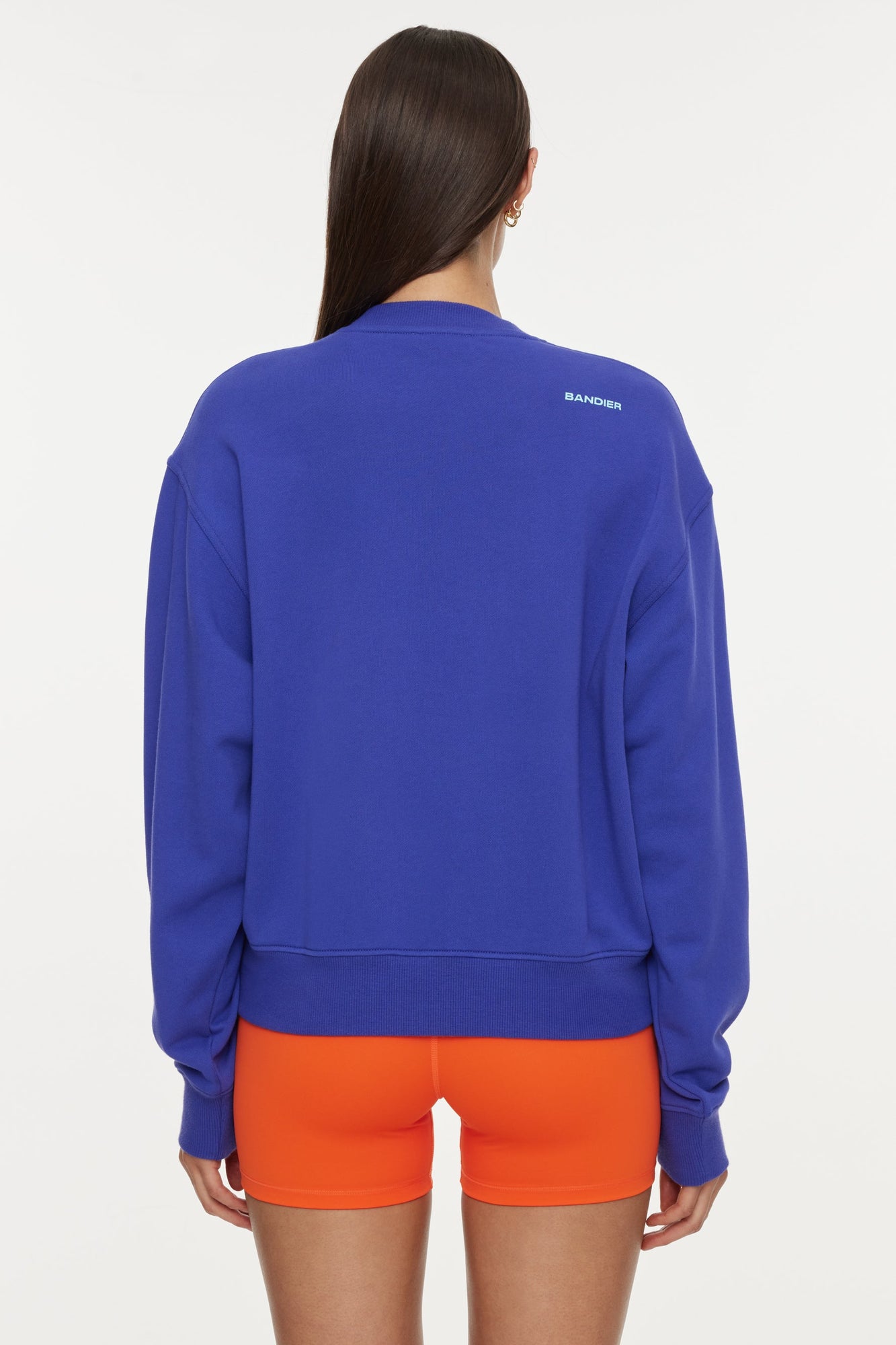 Model wears a royal blue crewneck sweatshirt with the lettering "Soleil Sport" across the chest with wavey lines below and "Bandier" in the bottom right corner. 