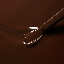 A close-up of a nail polish brush applying glossy brown nail polish, with smooth, reflective liquid covering the surface.