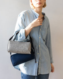 A person in a light denim shirt and jeans holds a graf lantz Campus Shoulder Bag made of two-tone fabric with black straps, standing against a plain background.