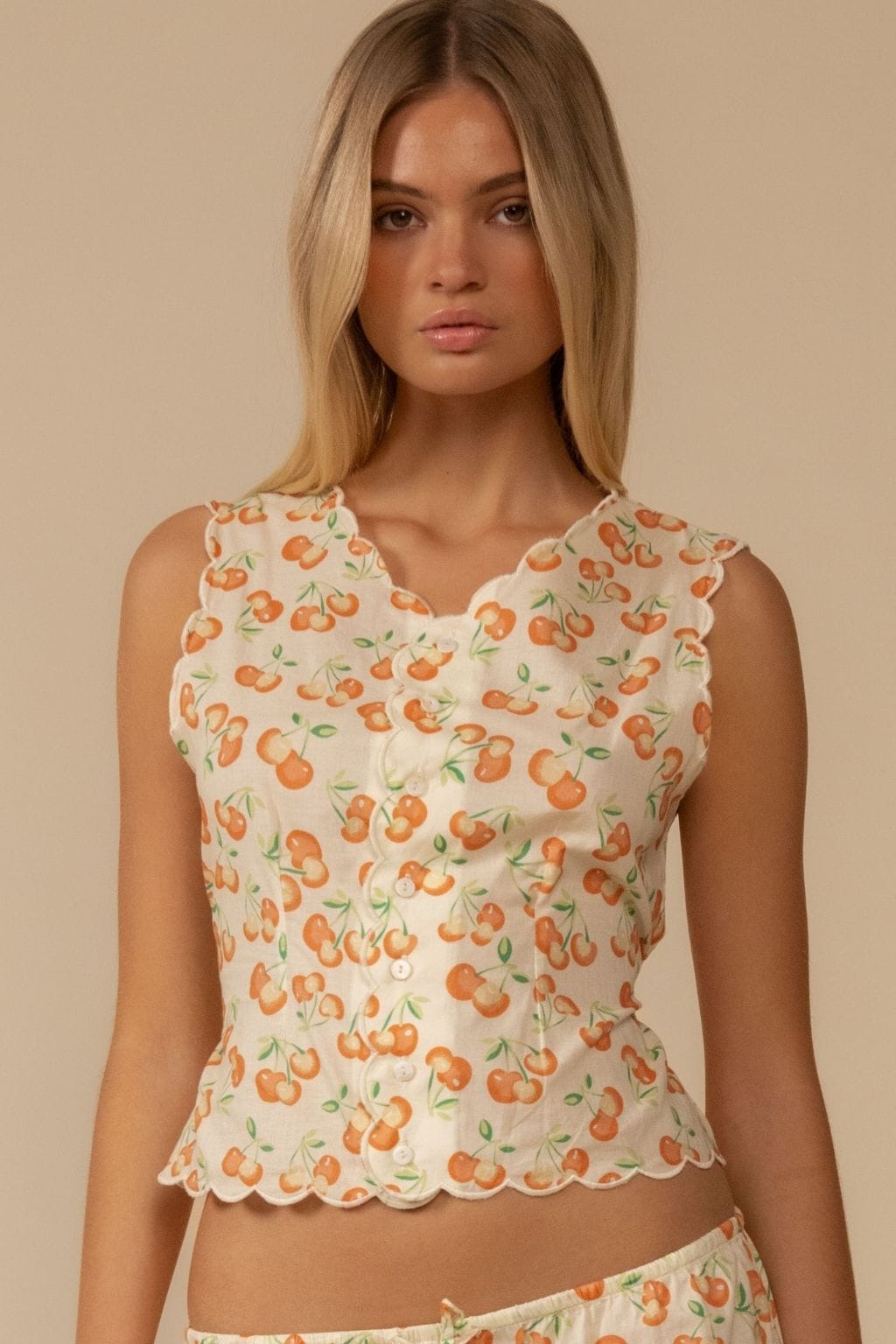 Sleeveless cream vest with orange cherry print and scalloped edges on neckline and hem.