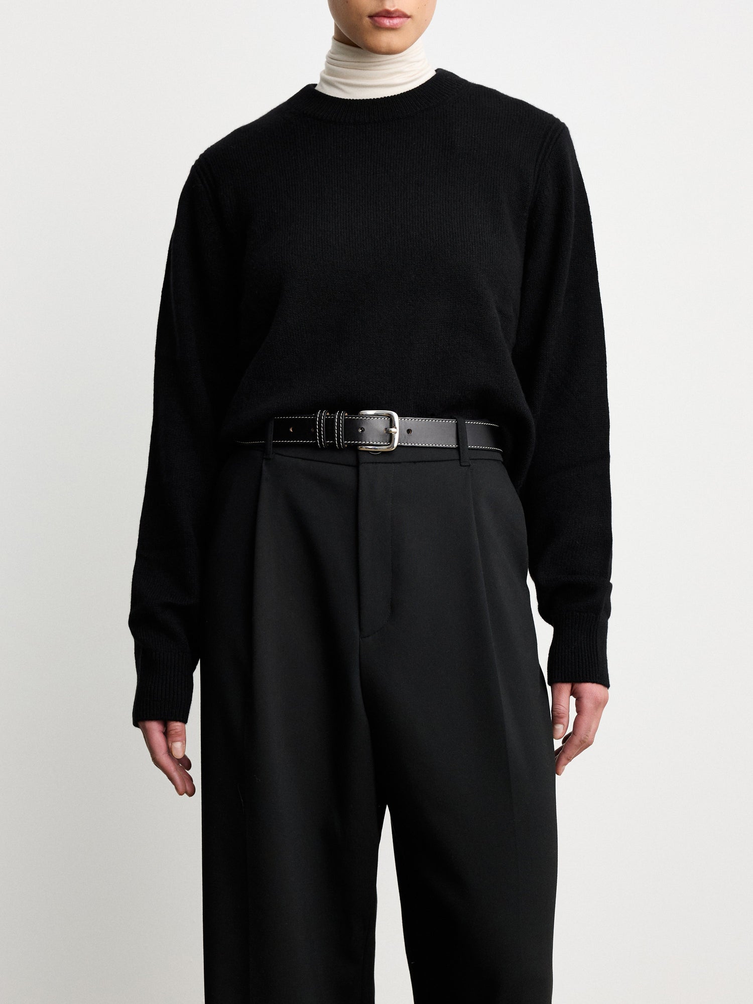 Model wearing Dehanche Louison black leather belt with white stitching and silver buckle, styled with black trousers and a black sweater