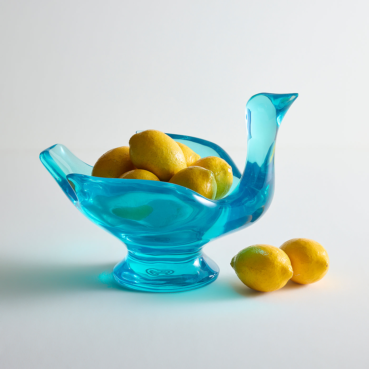 A translucent light blue glass bowl shaped like a bird, filled with yellow lemons, with two additional lemons placed beside it on a clean white studio background.